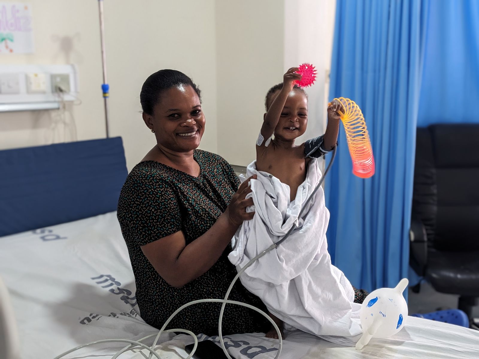 A mother smiles with her child, who waves a toy from a hospital bed